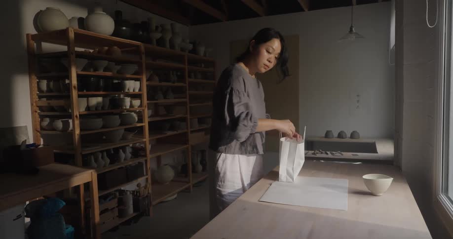 The video shows a woman in a pottery or ceramics studio carefully packaging an item on a wooden table near a windowshe places what appears to be tissue paper or wrapping into a small white paper bag with shelves of finished pottery visible in