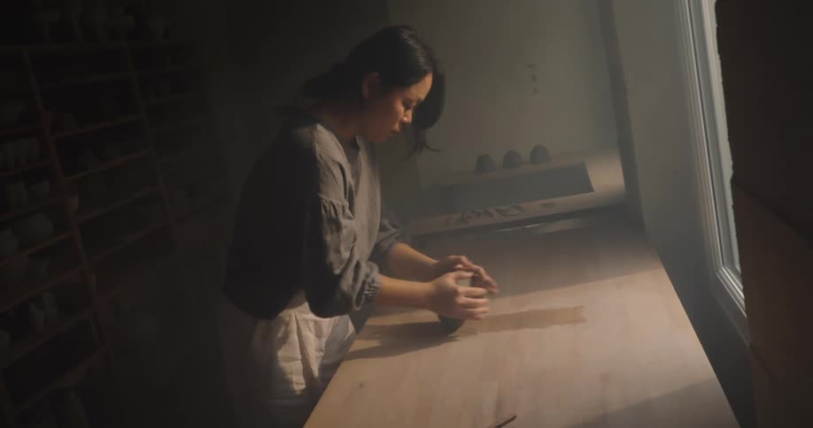 Woman kneads clay on a workbench beside a bright window