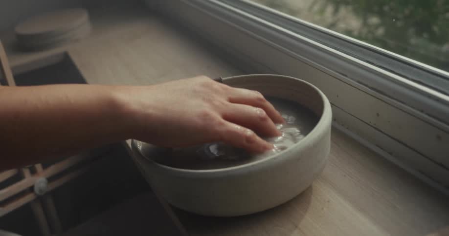 A person dips their hand into a bowl of water placed by the window rinsing it briefly