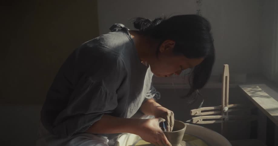 Close up of a young asian woman moulding pottery on a pottery wheel in a room during daytime