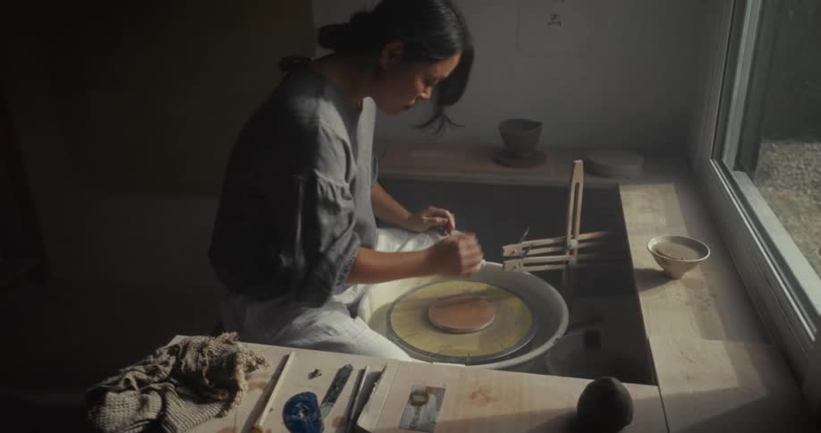 Woman works with clay on a pottery wheel near a bright window
