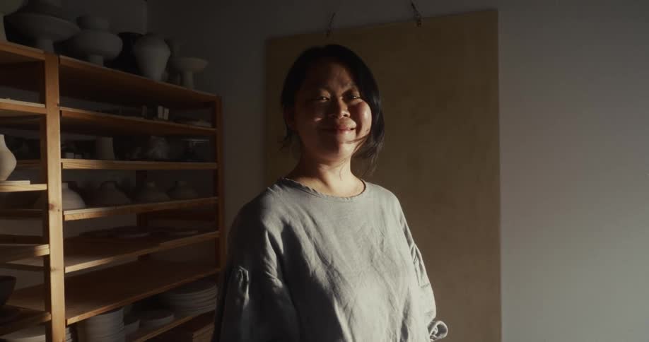 An asian woman standing in a room with shelves of ceramic objects on the left and a brightly lit wall on the right