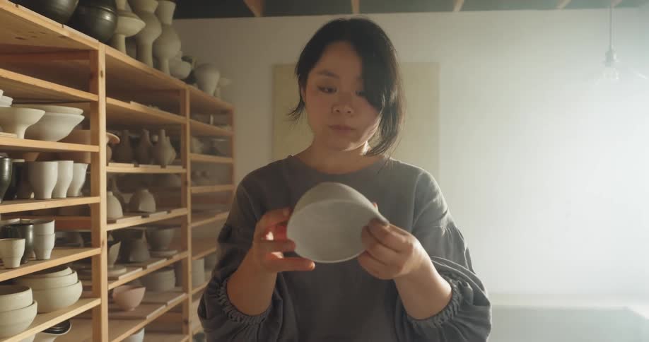 A young asian woman examining a ceramic bowl while standing in a pottery room