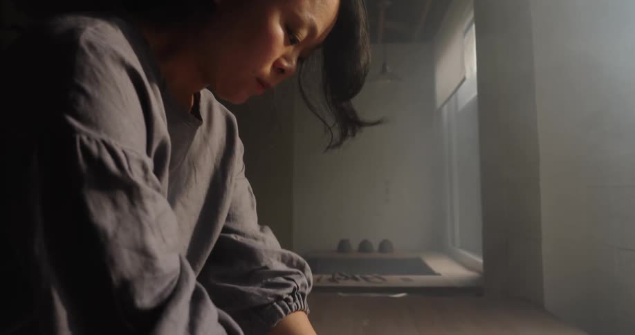 A closeup of a woman with dark hair standing over a wooden table bending over and using both hands to press and knead a clay on the surface