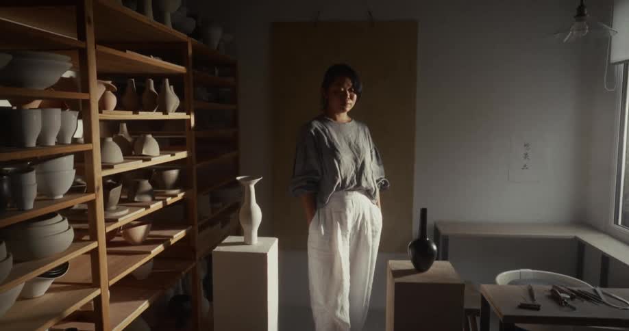 A young asian woman standing in between displayed ceramic pottery in a pottery room during daytime