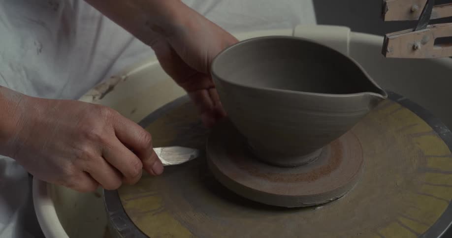 A persons hands using a thin metal tool to cut an unfired clay vessel with a spout shape from a small wheel bat then lifting the finished piece away from the pottery wheel