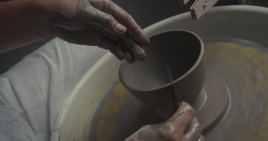 A persons hands holding a tool to cut the base of an unfired clay vessel that is spinning on a pottery wheel