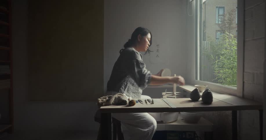 Person reaches for a bowl near the window in a ceramics studio with work tools on the table