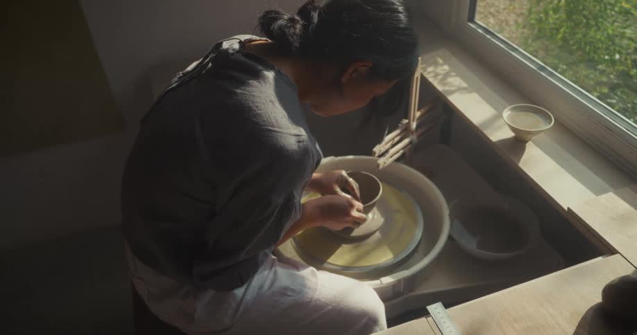 A woman is sitting at a clay wheel by a window using the wheel to sculpt a clay vase