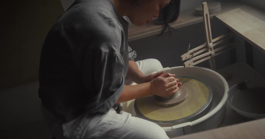 A person with dark hair tied back sitting at a potters wheel near a large window using both hands and forearms to firmly press and center a lump of clay on the spinning wheel head