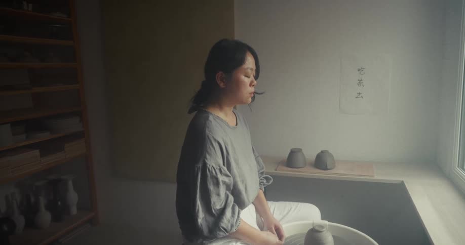 A woman sitting at a pottery wheel near a window with her eyes closed in concentration