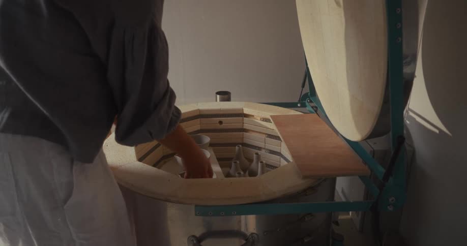 Close up of a young asian woman placing clay pottery in a kiln for firing in a room during daytime