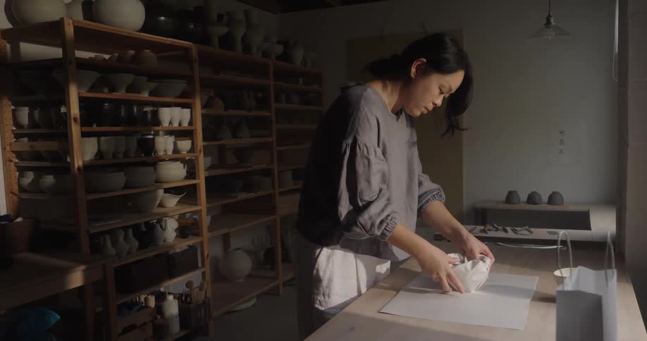 A young asian woman wraps a clay cup in paper and places it into a gift bag for delivery while in the studio at midday