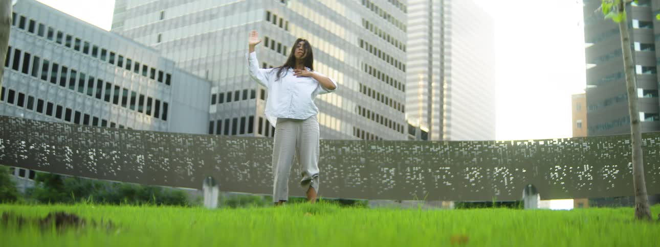 Woman dancing barefoot on a lawn surrounded by modern buildings