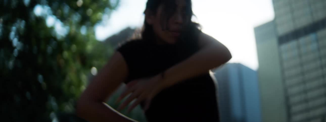 The silhouette of a hispanic woman with long hair moving their head and upper body while facing a bright sun flare and standing in front of outdoor foliage and buildings