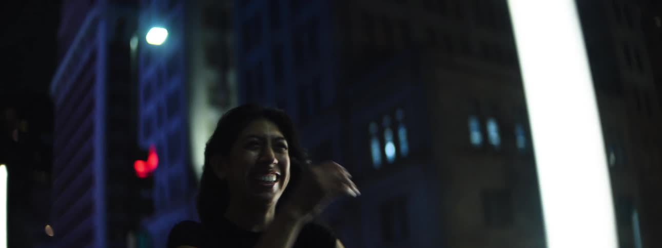 A hispanic woman with dark hair walking and smiling through a dark city street at night