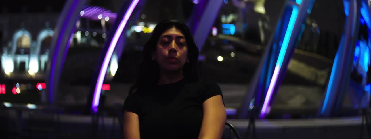 Woman sits alone at a table in a nighttime setting illuminated by colorful lights