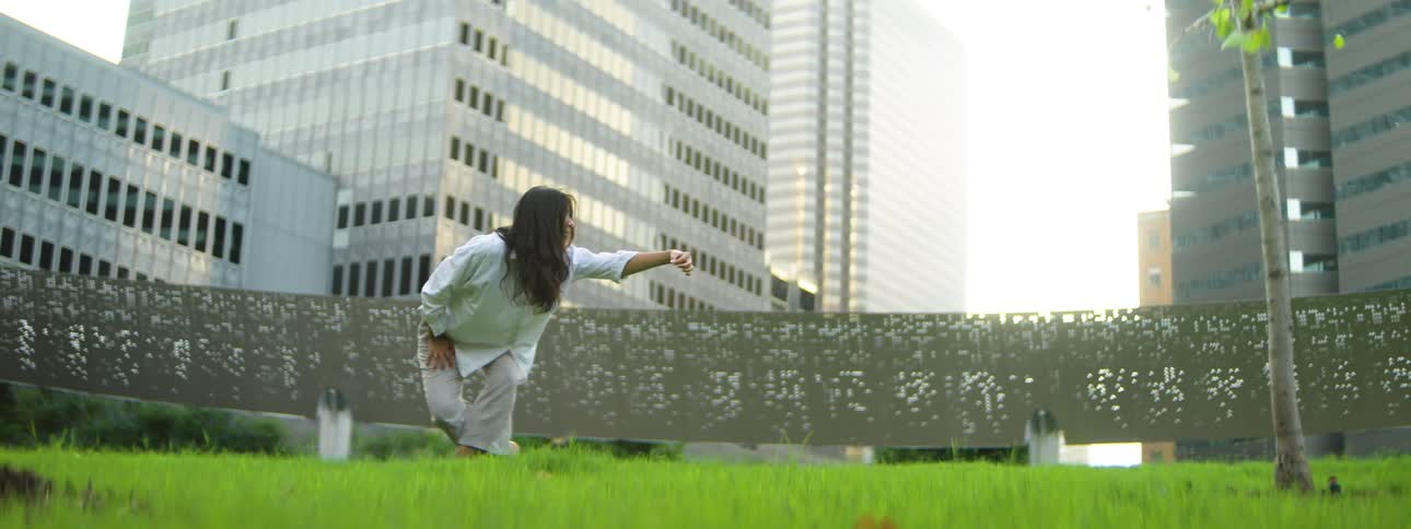 A hispanic woman with long dark hair standing on a patch of grass in an urban area with tall buildings in the background