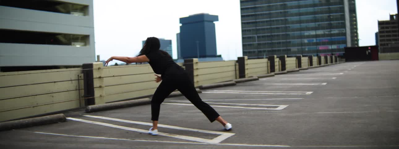A woman with dark hair dressed in black clothing performs dance movements on a paved surface marked with white lines backed by a row of buildings and a cityscape under a light sky