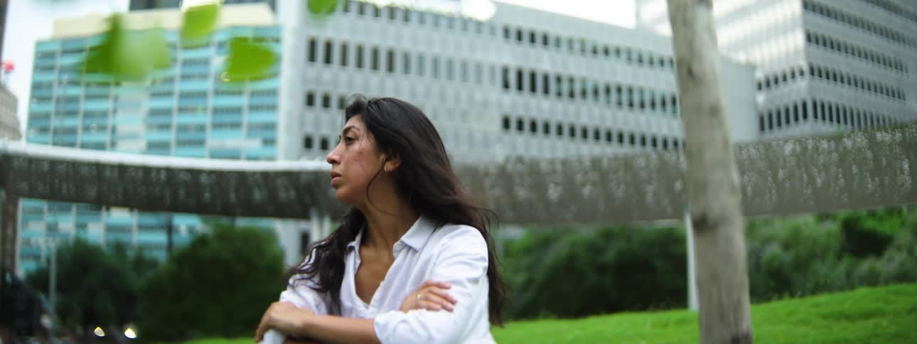 A woman in a white shirt sits on green grass gazing thoughtfully to the side with a modern office building behind her
