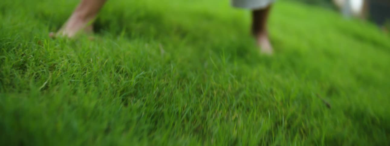 A woman with exposed lower legs wearing light fabric clothing walks and bends while barefoot on a thick carpet of green grass