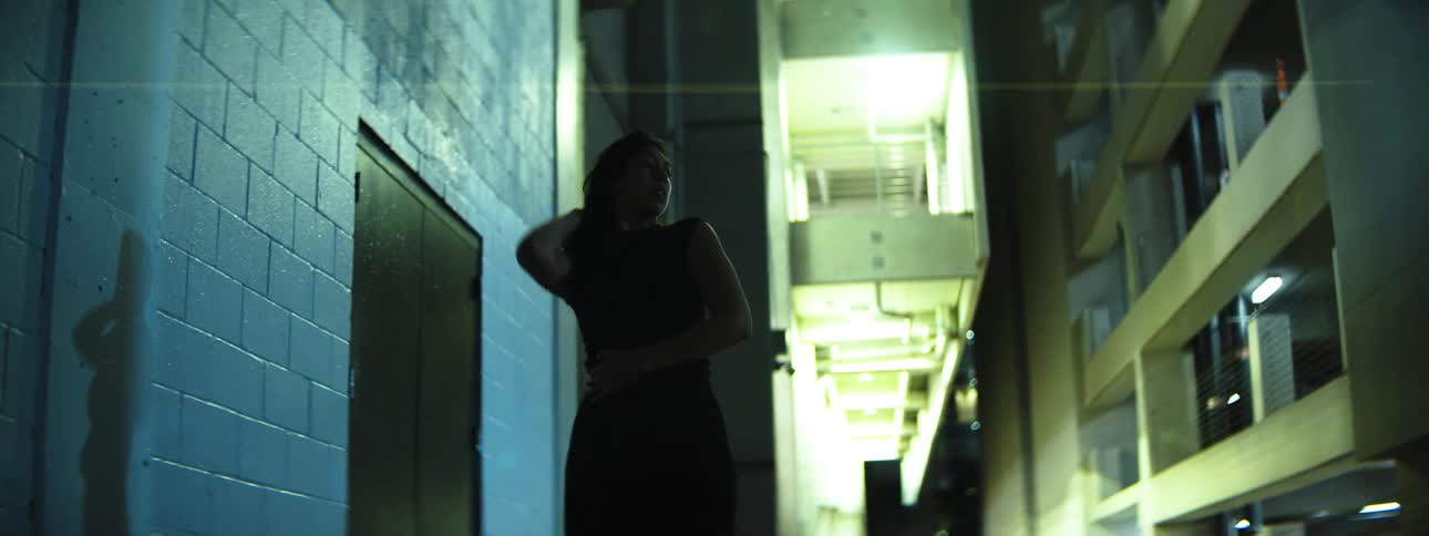 A hispanic woman with long hair dressed in dark clothing performs fluid dance movements against a textured blue wall in a very dark narrow outdoor passage