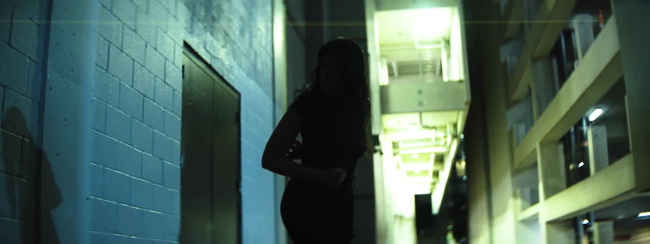 A hispanic woman with long dark hair wearing a dark garment stands and performs small stylized movements in profile appearing as a dark silhouette against a brightly illuminated background structure at the end of a narrow dark passag