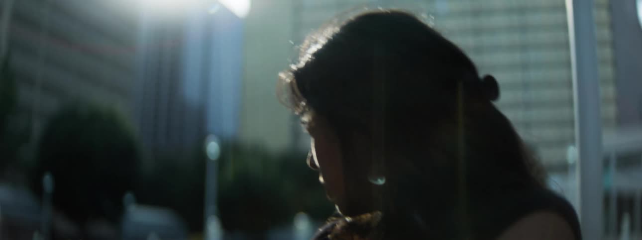 A woman in dark clothing sits outdoors against a city backdrop her face half in shadow as she looks sideways with a pensive guarded expression