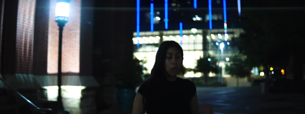 A woman leaving a building facade and looking around in a dark outdoor area near steps and a street lamp with a brightly lit building in the distant background