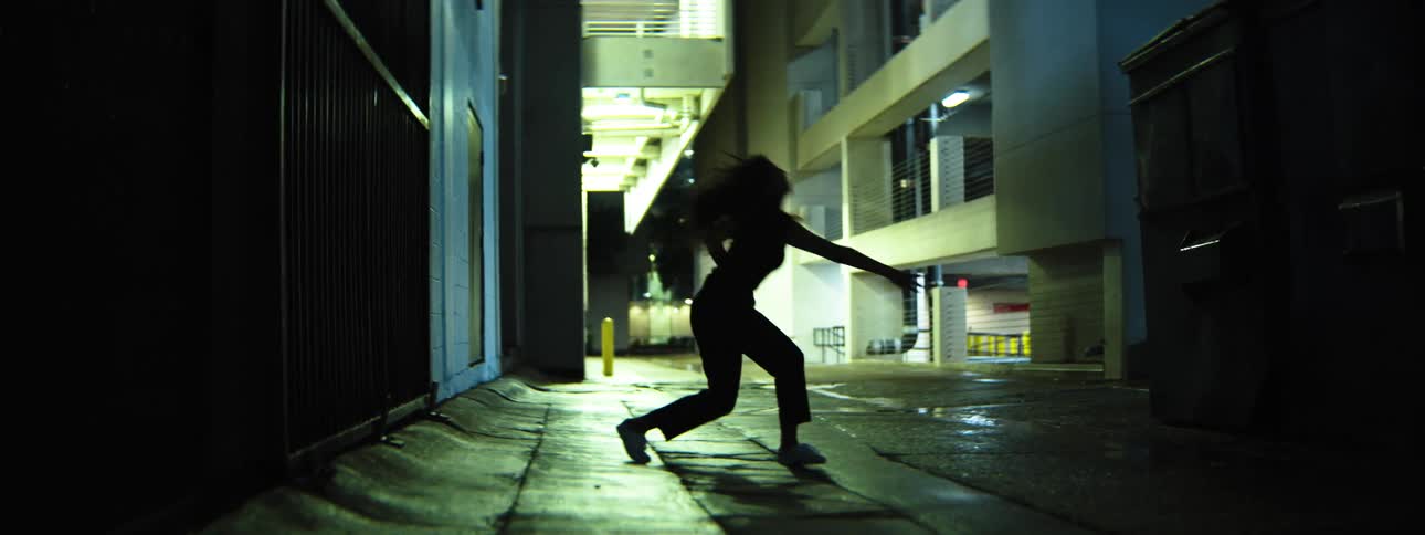 Awoman with long dark hair dressed in dark clothing performs dance movements with sweeping arm and leg gestures