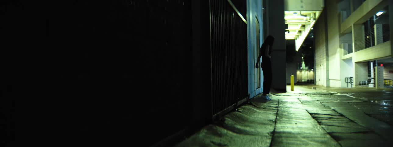 A woman stand still facing a tall gate in a wet urban alleyway at nigh