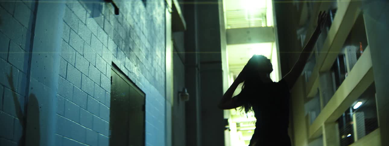 A woman in shadow stretches her arms upward in a dimly lit alleyway illuminated by harsh overhead lights