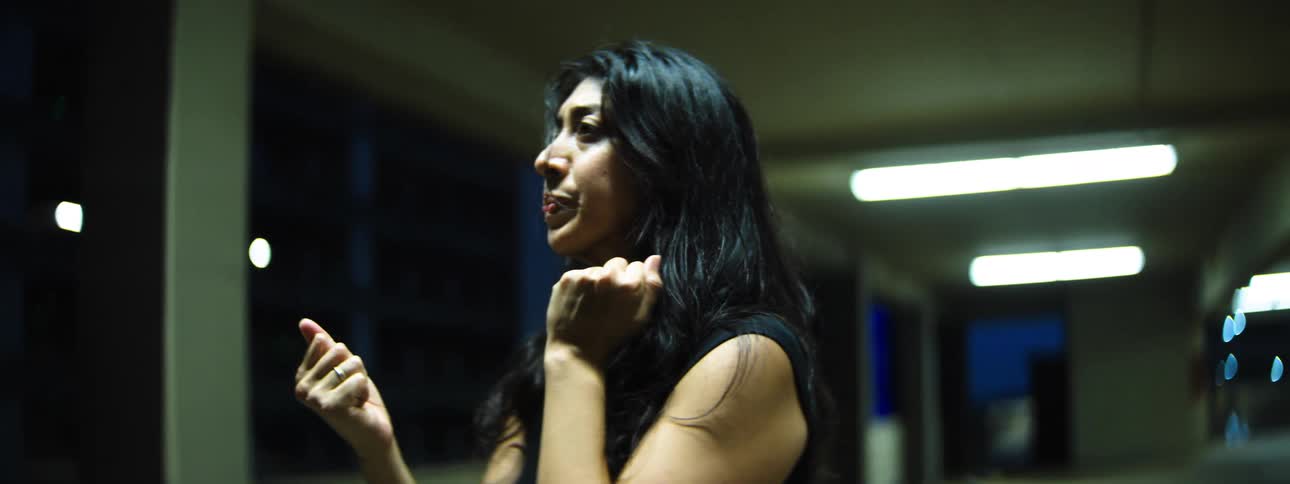 A woman under fluorescent lights holds her clenched hands near her face looking serious and determined