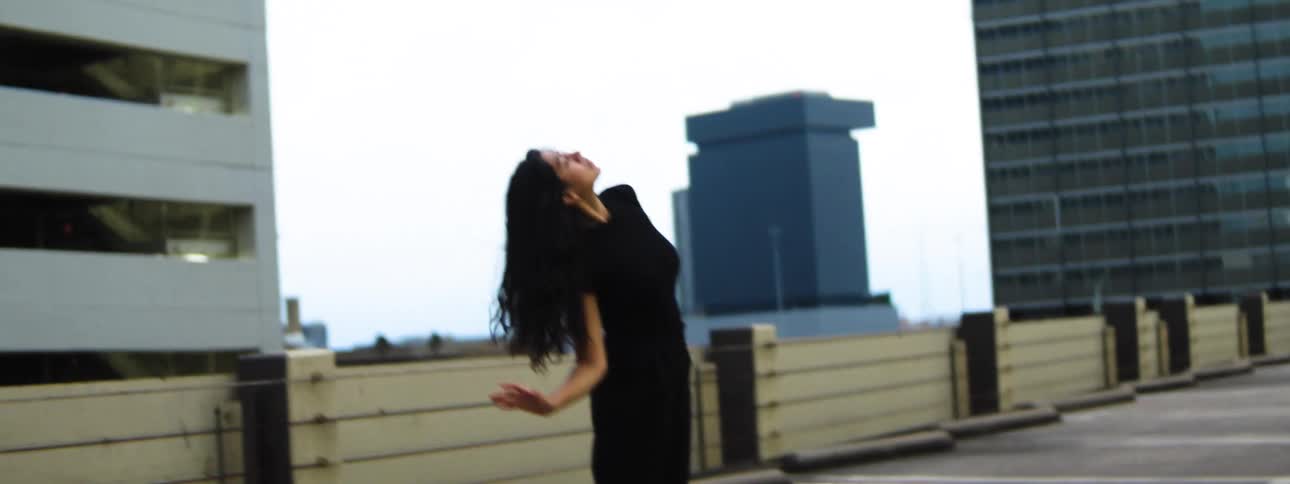 A woman in a dark outfit dances atop the roof of a city parking structure