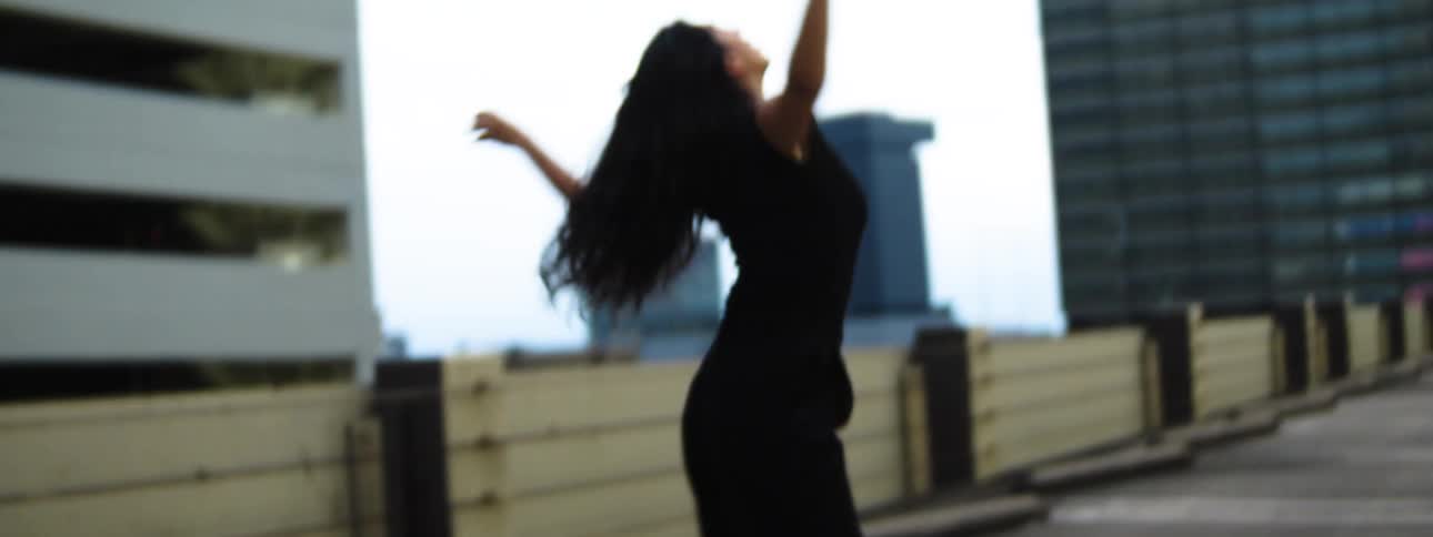A woman in black stands on a rooftop parking deck with arms flung wide smiling joyfully against tall city buildings