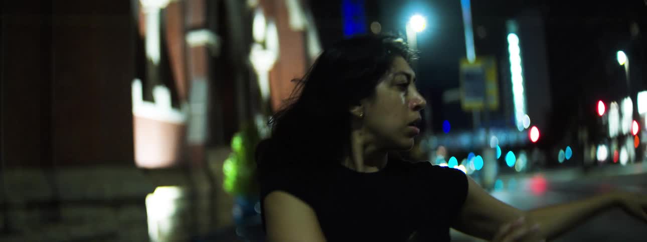 A woman with long dark hair performs expressive dance movements on a brightly lit city street at night