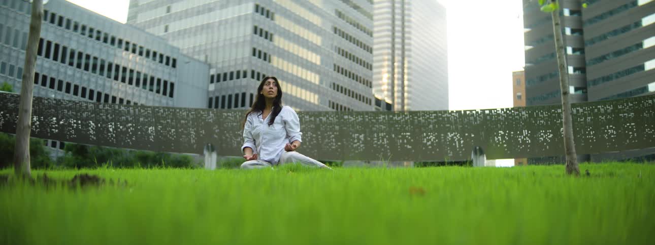 Female in an oversized white sweatshirt tilts her head while sitting on grass surrounded by buildings