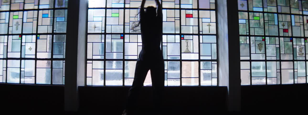 A woman silhouetted before a vast stainedglass wall holds her arms out appearing calm and contemplative
