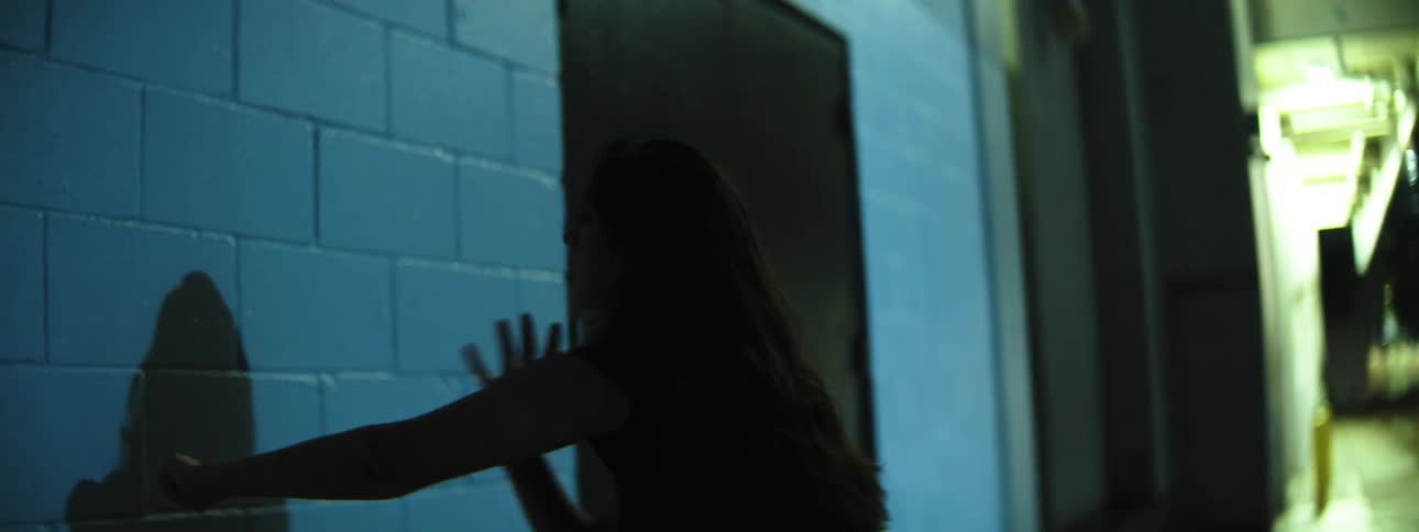 A woman in a dim hallway extends her arm toward a blue brick wall looking focused and wary
