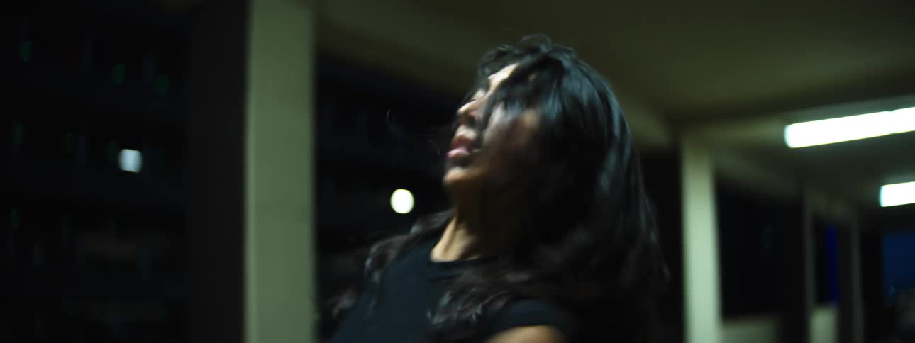A hispanic woman with long dark hair and wearing dark clothing moves their head and arms with intensity in a corridor at night