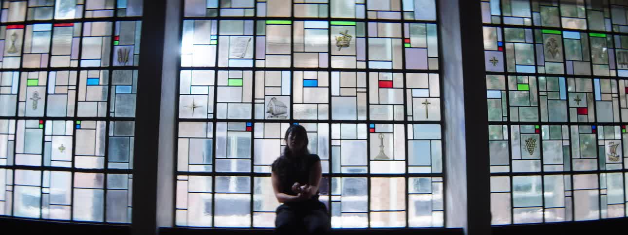 A woman stands silhouetted against stained glass windows moving her arms in the air before sitting