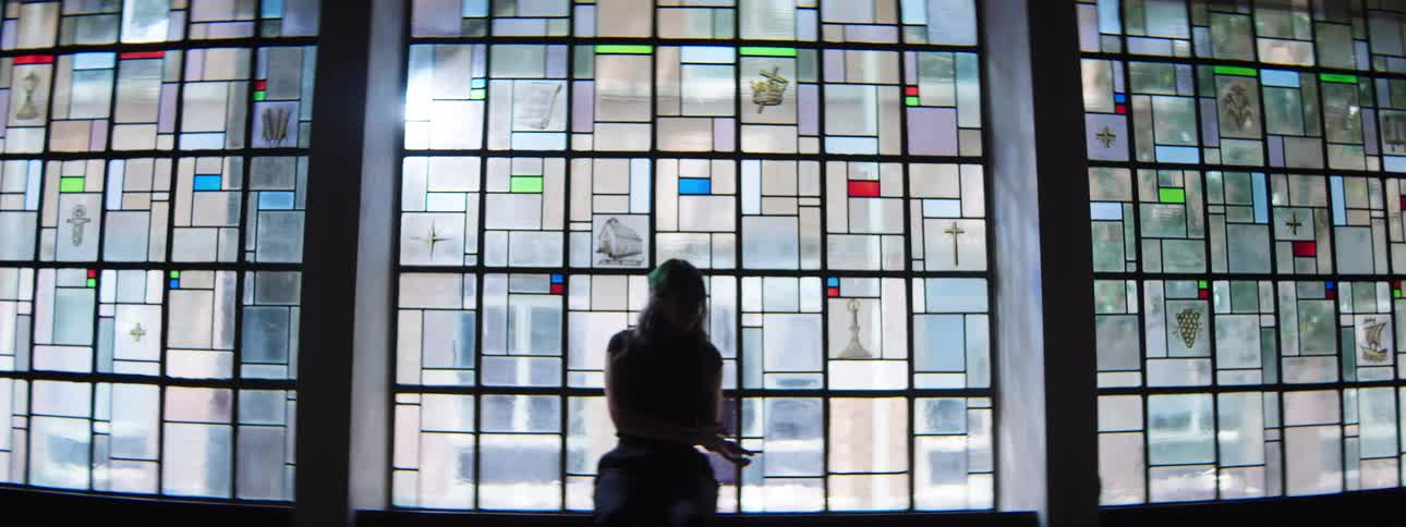 A hispanic woman with long hair dressed in dark clothing dances in a large brightly illuminated window composed of a geometric pattern of clear and colored glass