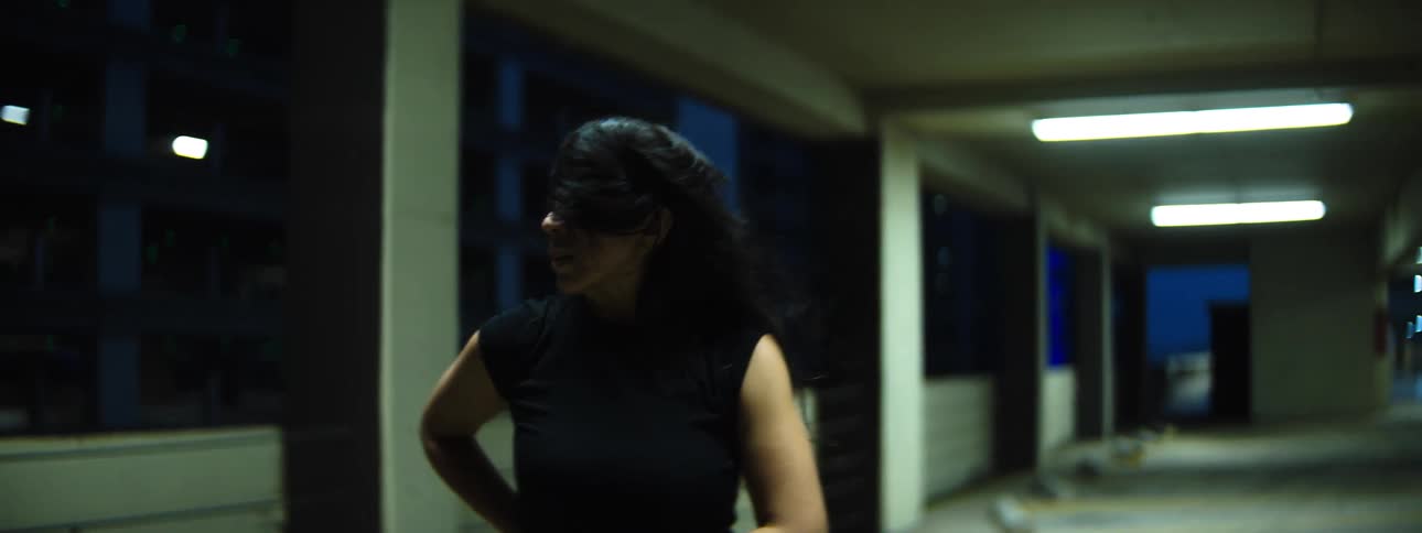 Female dancer in a dim parking garage extends her arm with fierce concentration hair tousled and neon lights blurred behind
