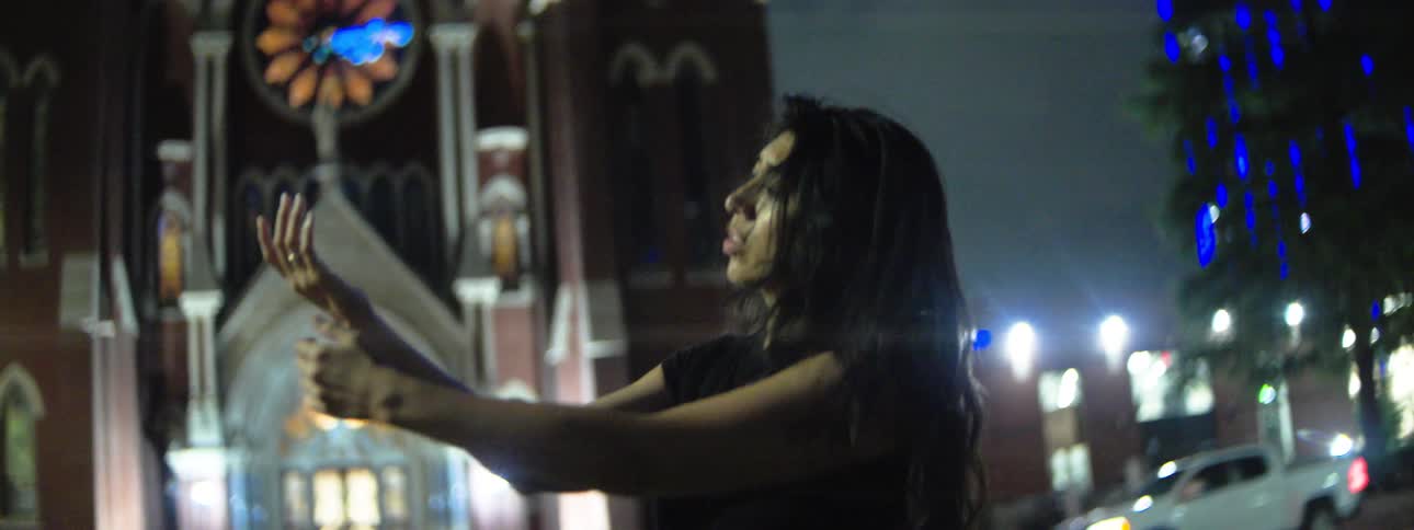 A woman stands before a lit church at night one hand on her chest and the other raised looking sad