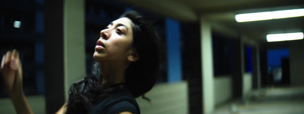 A dancer with long black hair performs expressive fluid movements with her arms and body in a dimly lit urban tunnel