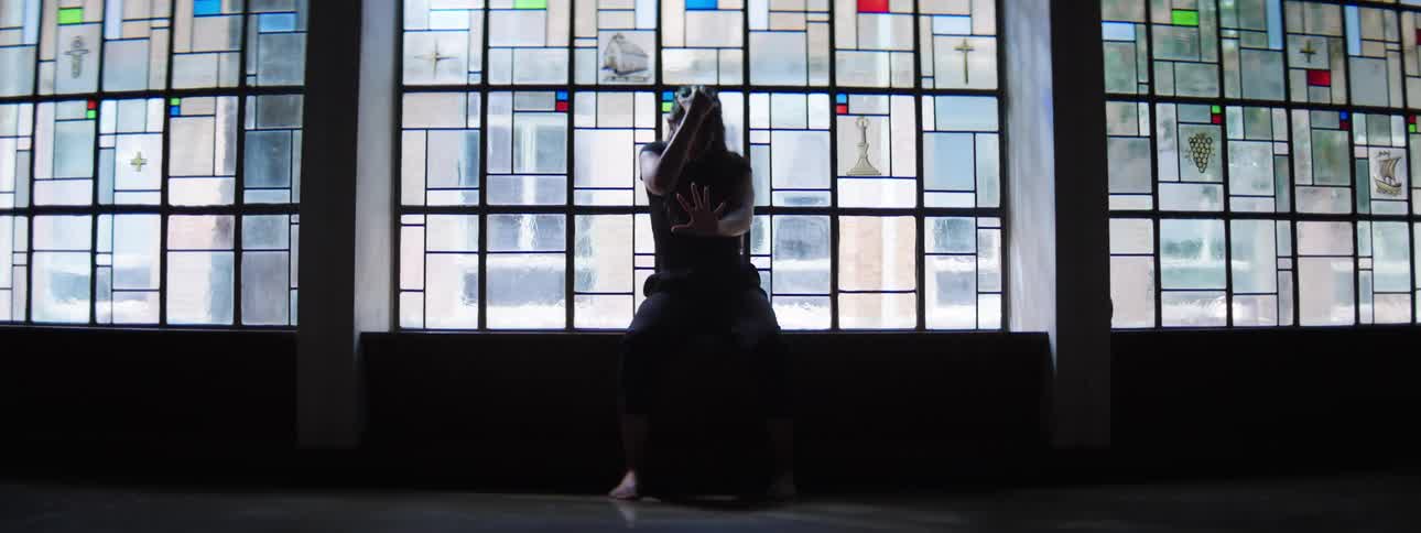 A hispanic woman performs a series of fluid and grounded dance movements moving between standing on the floor and sitting on a window ledge in an indoor setting