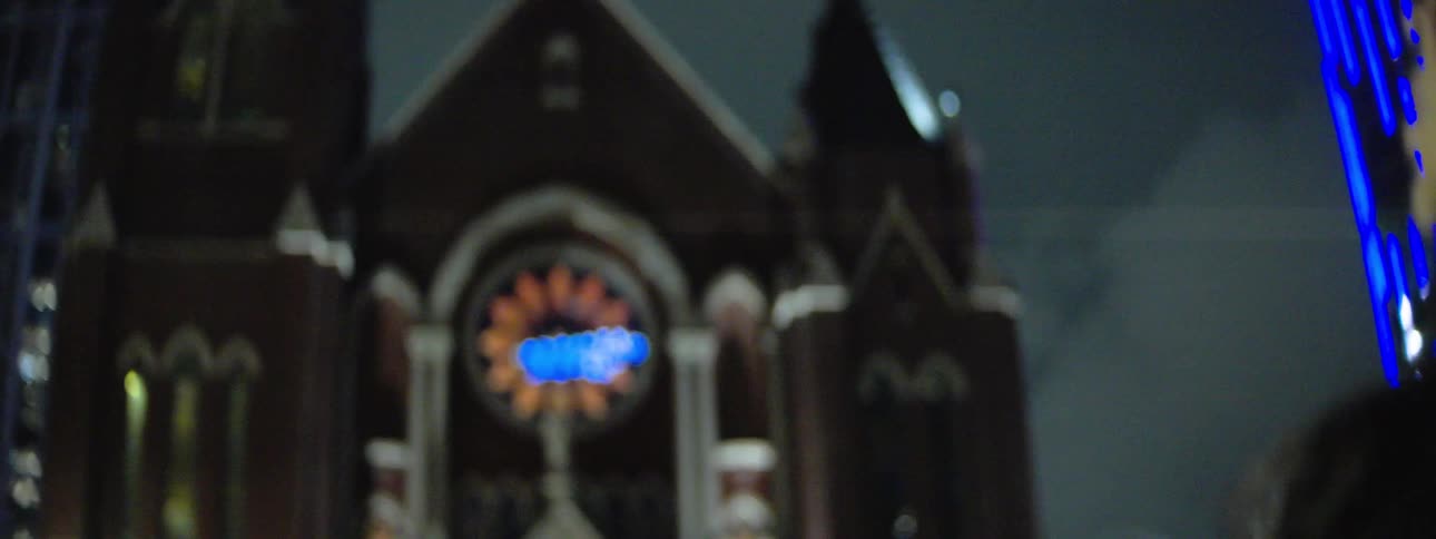 A hispanic woman with long dark hair raising their arms and moving their hands with an emotional expression transitions to a view from behind the person as they stand facing the illuminated facade of a church building at night