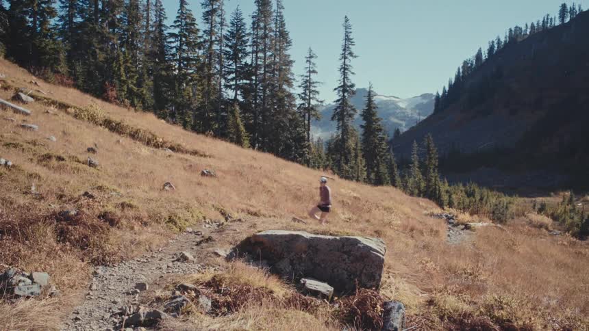 A woman jogs along a mountain trail through trees and grassy slopes