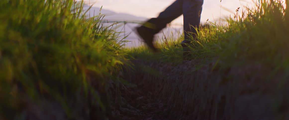 A low groundlevel view captures a persons legs wearing dark pants and work boots quickly stepping over a small dirt ditch between two patches of bright green grass with an irrigation sprinkler visible in the background at sunset