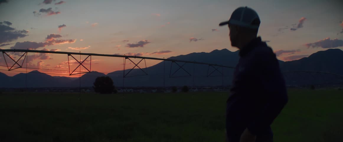 A man facing away from the camera and wearing a hat stands in silhouette on a large field with a centerpivot irrigation system structure visible on the left set against a background of mountains and a sunset